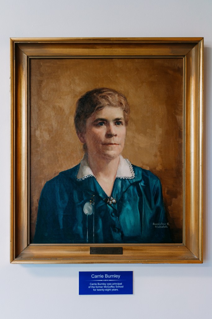 A portrait of a woman in a blue blouse with a gold frame hangs on a white wall. Beneath the portrait is a sign that says, "Carrie Burnley."