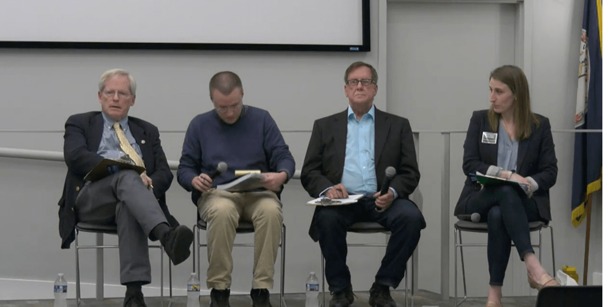 Four people sit in chairs in a row with a white wall and flag behind them.
