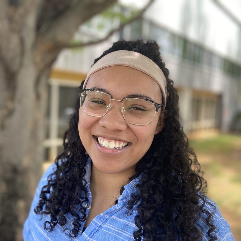 A woman with curly hair and a headband smiles for camera with a tree and building behind her