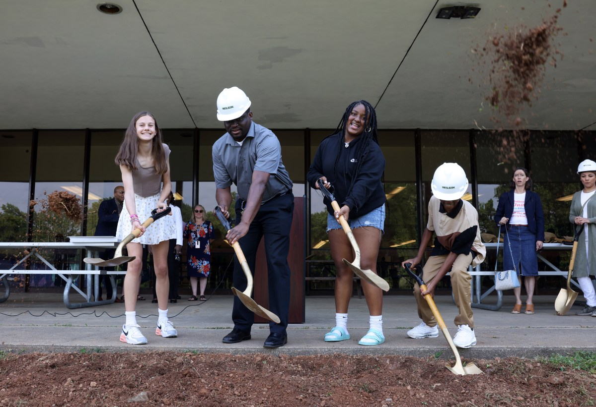 A man in a hard hat is surrounded by three teenagers. Each person has a shovel and is digging into dirt. They stand in front of an old, brick building.