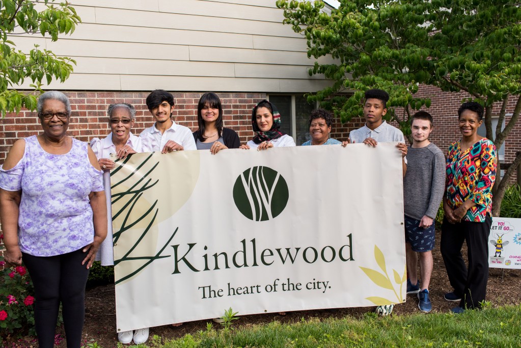Nine people of varying ages and races stand, smiling, in a line behind a banner. The banner reads "Kindlewood" in large text, with "The heart of the city" in smaller letters beneath it. 