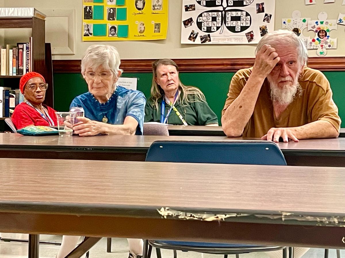 Four people sit at long tables in a common area looking upset.