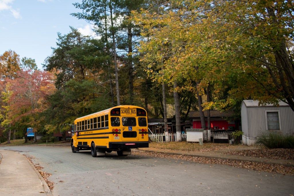 A school bus is pictured from behind in a trailer park surrounded by trees with changing leaves.