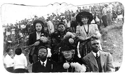 In the foreground, a group of six Black people — three women, two men — wearing fine clothing and posing for the camera. Behind them, a gathering of dozens of people, also dressed up.