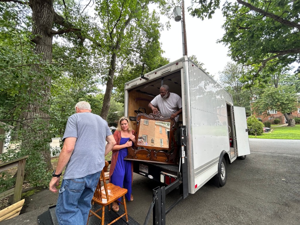 A man hoists a piece of furniture out of a white truck while another man (back to camera) pushes forward a chair. A woman in a blue jumpsuit stands between, helping.
