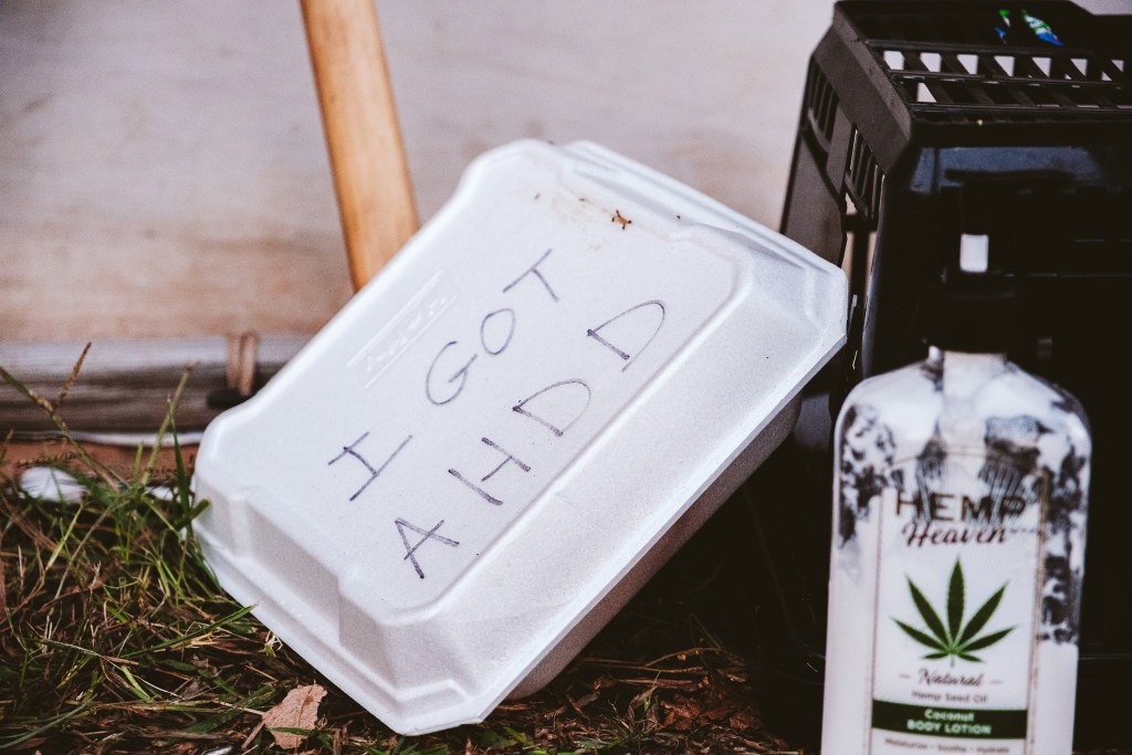 A styrofoam food takeout container sits on the ground outside a tent. The letters "I GOT AHDD" are written on the top in black marker.
