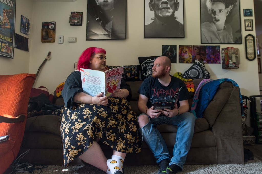 Two people sit on a sofa with many pop culture posters and pictures on the wall behind them. They are looking at each other, smiling.