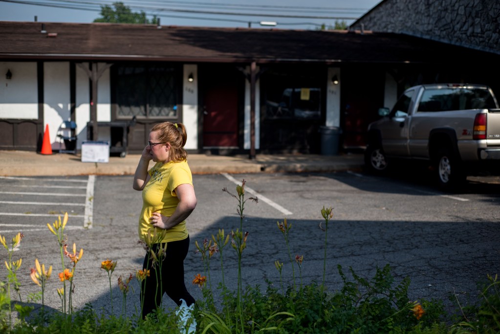 A woman in a yellow t-shirt talks on the phone in a parking lot, in front of a one-story motel, with a truck parked in front.
