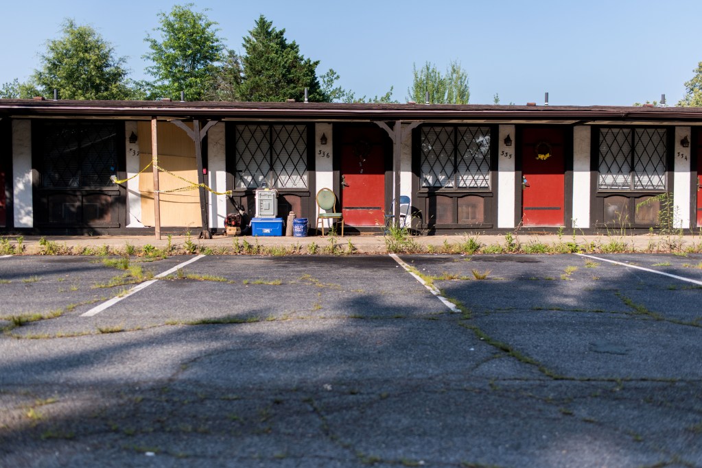 The front of a motel-like building, with wood panels and support structures holding up the crumbling awning, A cooler and chair are among peoples belongings that sit in from the doors.