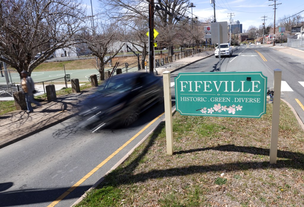A sign in the median of a road reads, "Fifeville" and "Historic, Green, Diverse." Two cars drive by the sign, with a basketball court to one side.