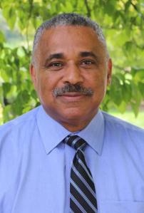 A man in a tie smiling for camera, leaves behind him.
