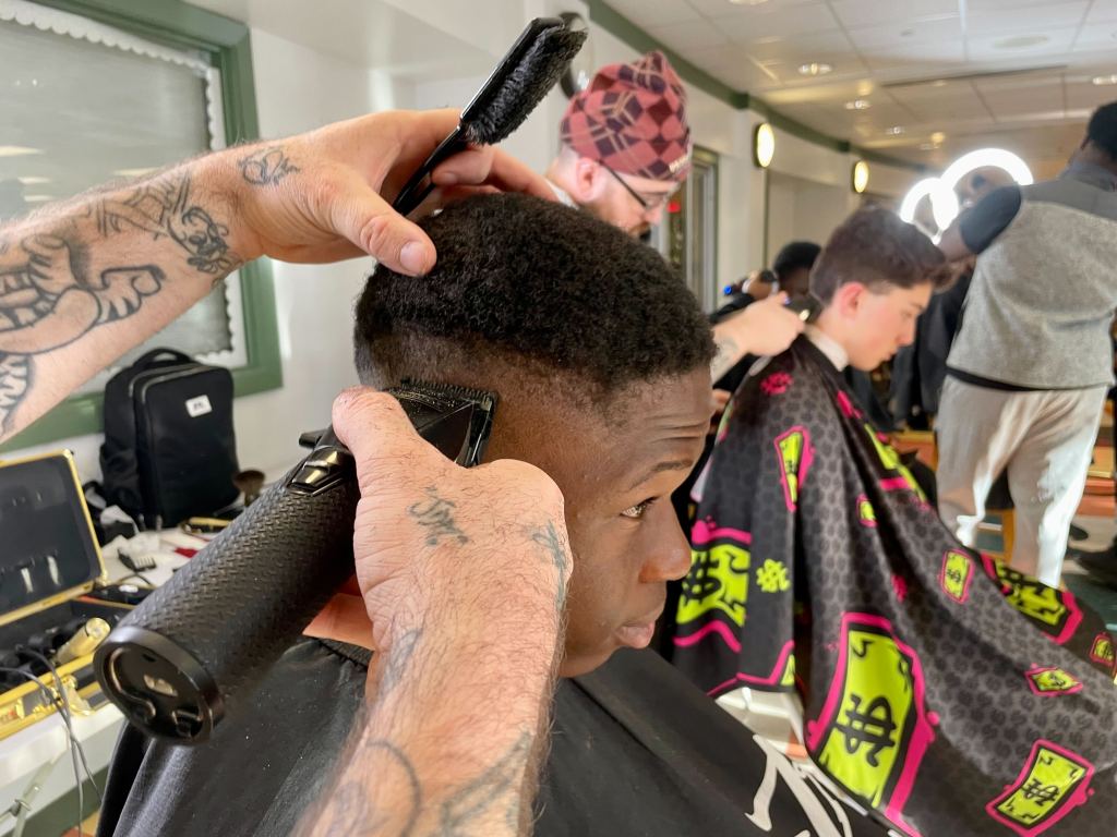Two teens sit in chairs getting their hair cut by two men in a room full of people.