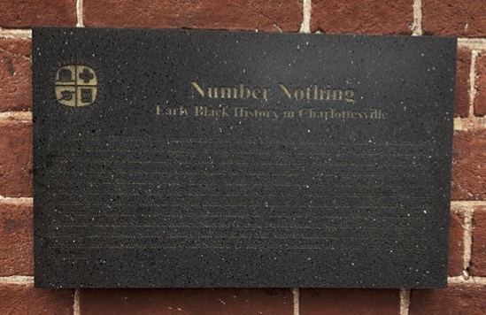 A photograph of a black stone plaque on a brick wall. The words carved into the stone are nearly impossible to read — they are very small and the gold inlay is faded. The crest of the City of Charlottesville is in the top left corner.