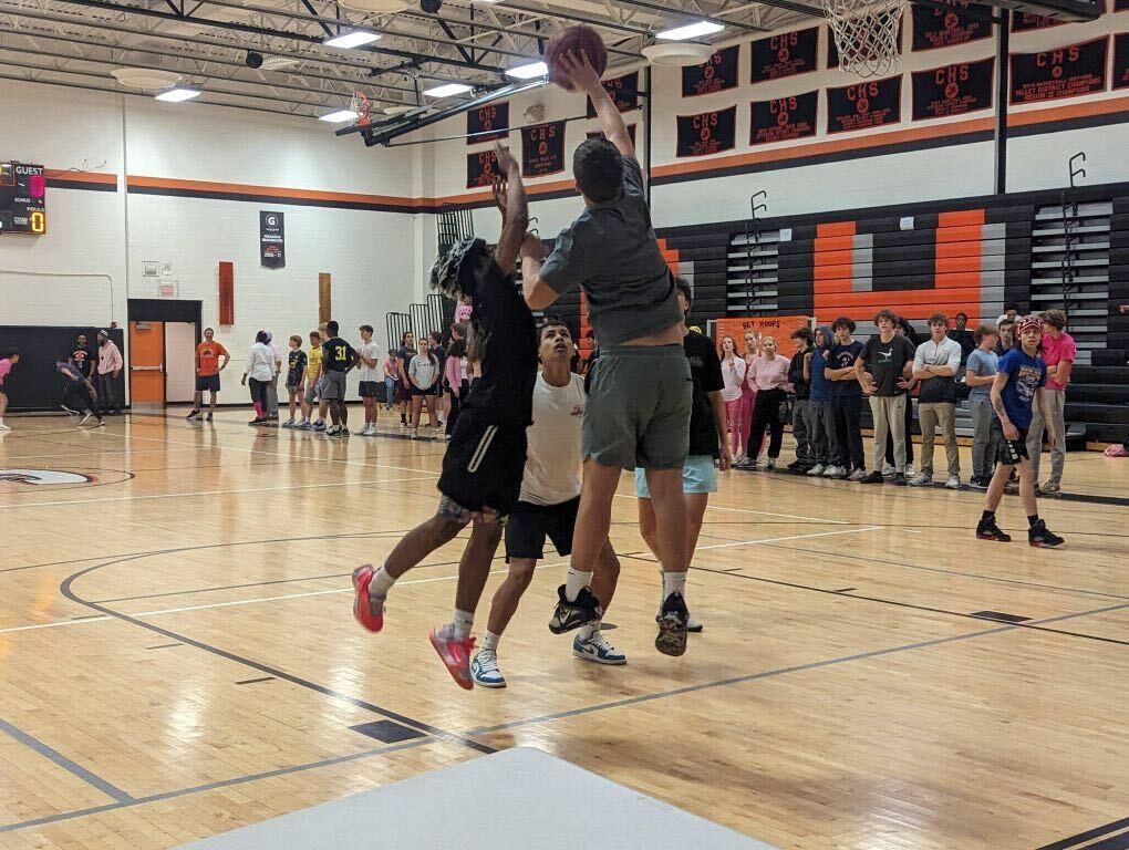 Teens play basketball on an indoor court. Several are jumping for the ball near the hoop.