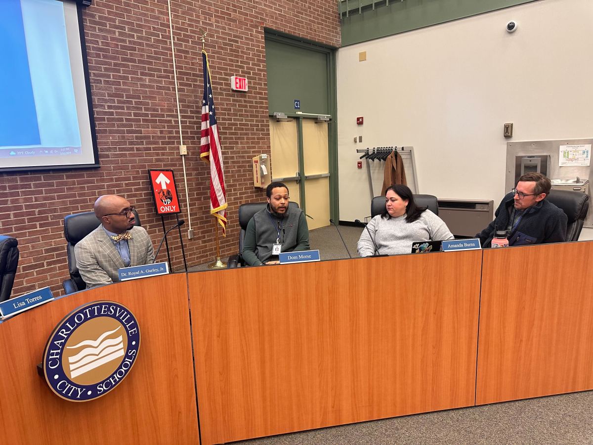 Four people talk behind a long desk, with a round logo to one side that reads "Charlottesville City Schools."