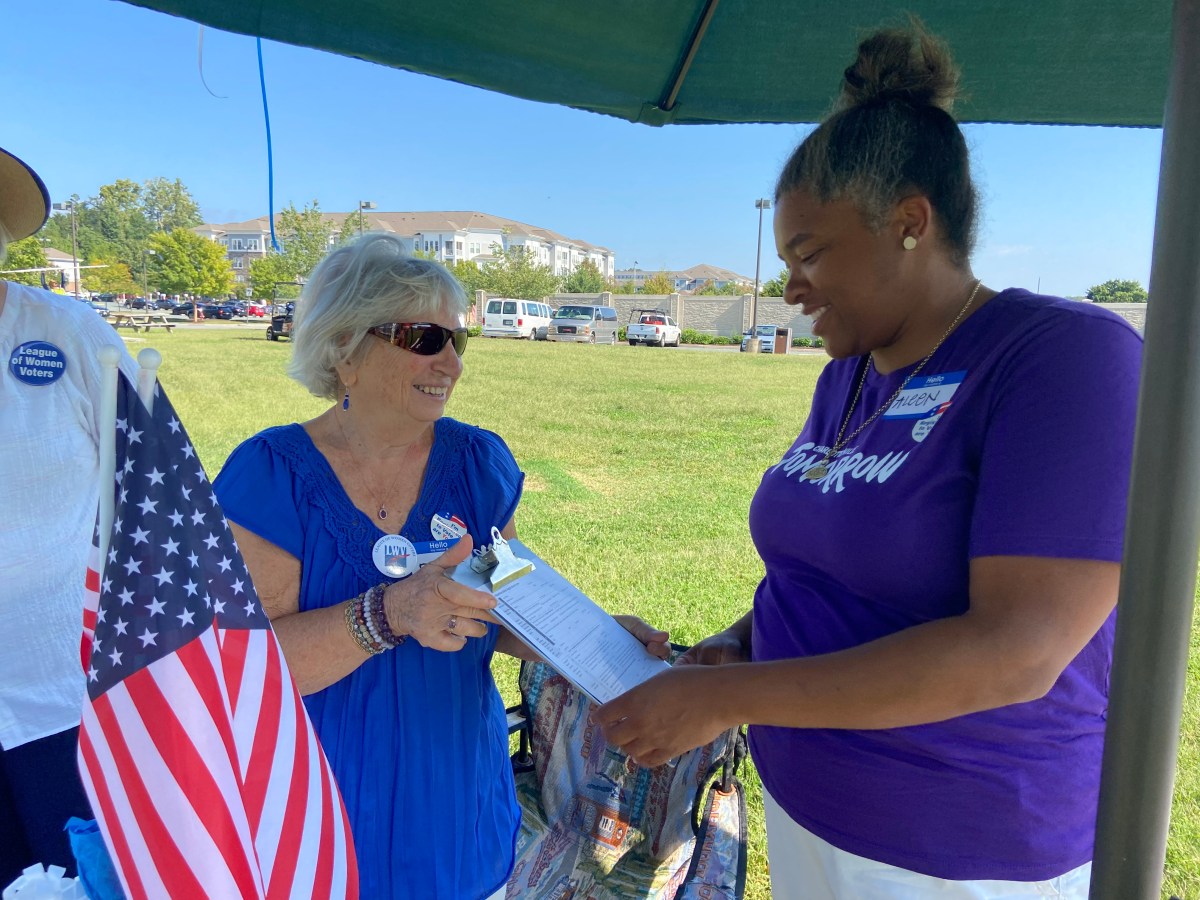Woman in purple shirt holds clipboard that she is handing to another woman. An American flag is in the foreground and green lawn behind.
