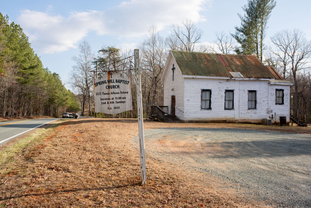 A one-story building with a cross on the front, next to a country road. A worn sign reads, "Spring Hill Baptist Church. Est 1803"