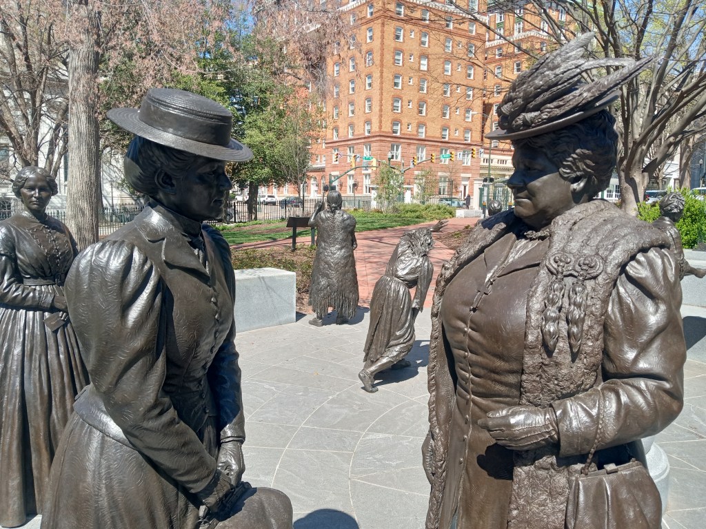 Two statues of women in skirts and hats face each other, with other life-size statues behind them.