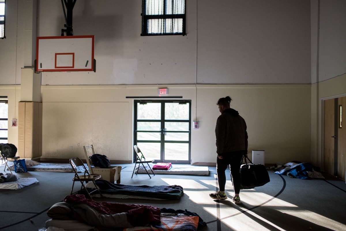 A person holding a duffel bag in a gym that has folding chairs and cots on the ground, with a basketball board on the wall and light streaming in.