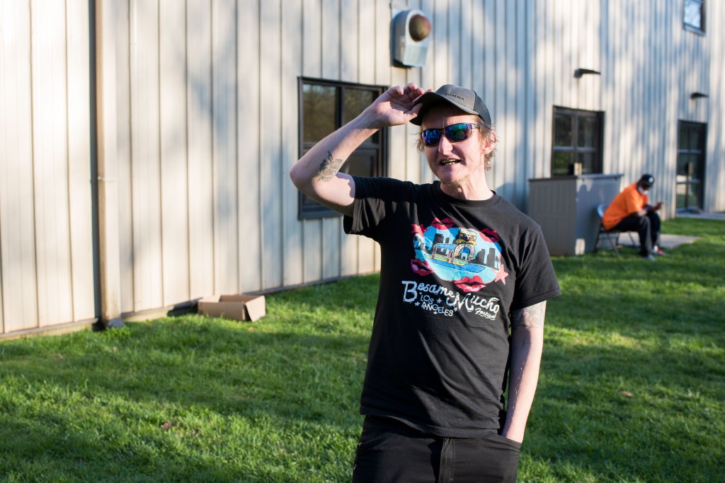 A man in a graphic t-shirt and baseball cap stands on grass in front of a building, holding his cap in the sun.