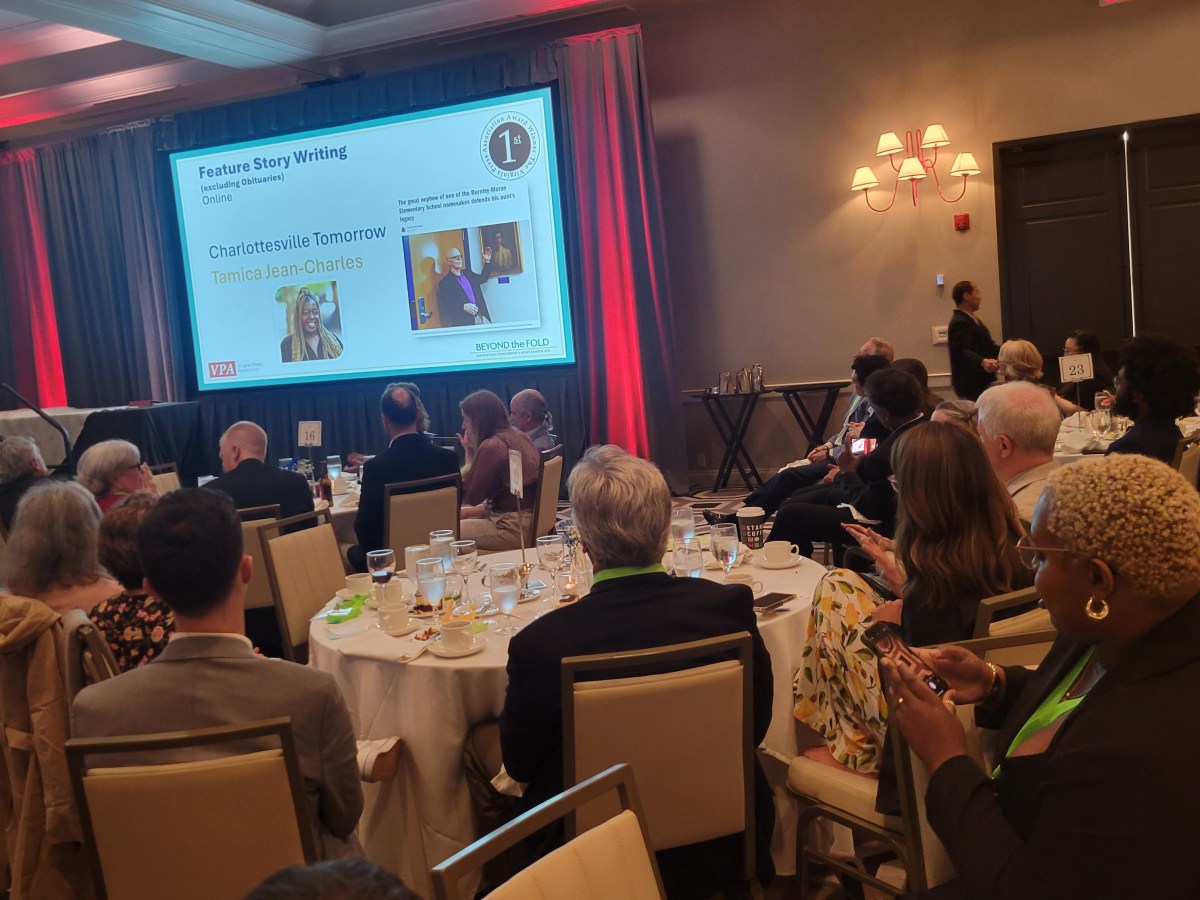A screen shows a person smiling with the words "Feature story writing" and the badge "First." The text says "Charlottesville Tomorrow" and "Tamica Jean-Charles," with people seated at dinner tables in the foreground.