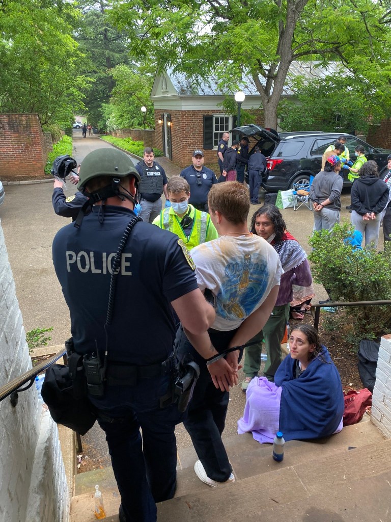 Police officer lead a person in a t-shirt, hands tied behind his back, dow a stairwell, while others look on.