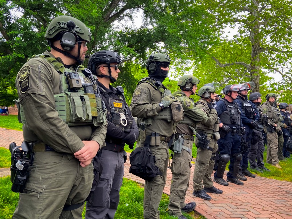 A line of officers in heavy gear, including weapons, masks, helmets with face shielfd and guns on a law with a brick walkway and leafy trees behind them.
