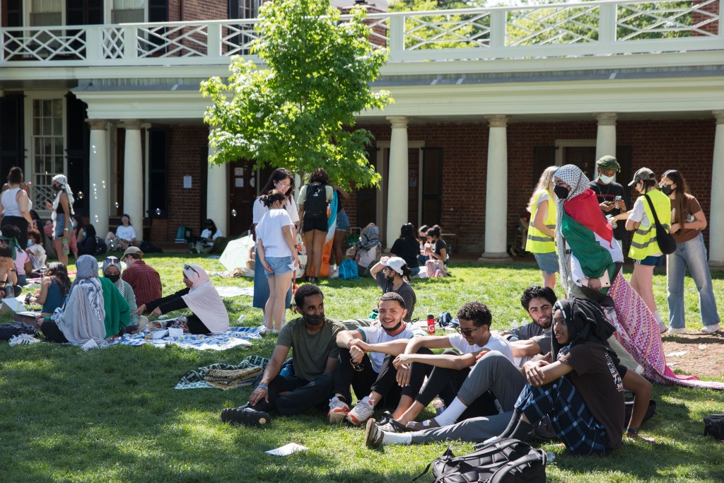 Dozens of people mingle in an outdoor grassy area with a brick building in the background.