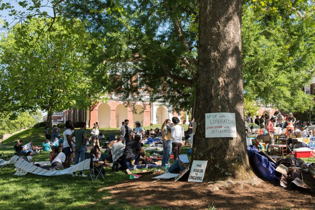 Dozens of people gather in the shade of a large tree, many have chairs and coolers. On the tree truck is a sign that reads: "up up with liberation, down down with occupation."
