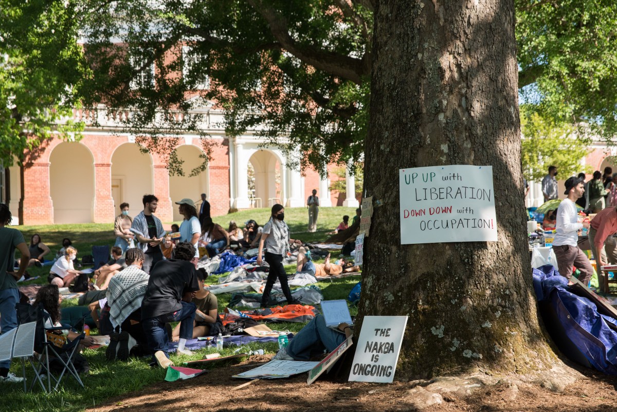 Dozens of people gather in the shade of a large tree. Attached to the tree is a sign that reads, "up up with liberation, down down with occupation."