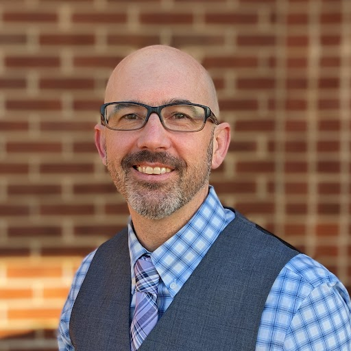 A person smiles for a portrait in front of a brick wall.