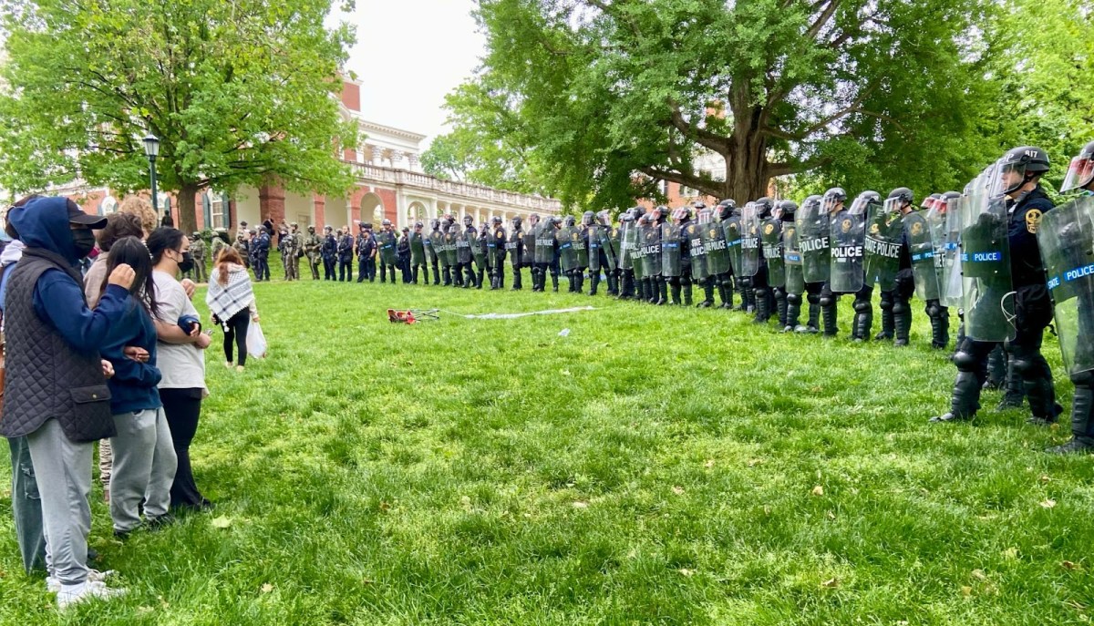 On a grassy area, a line of dozens of police officers in riot gear holding shields face off against people dressed in civilian clothing.