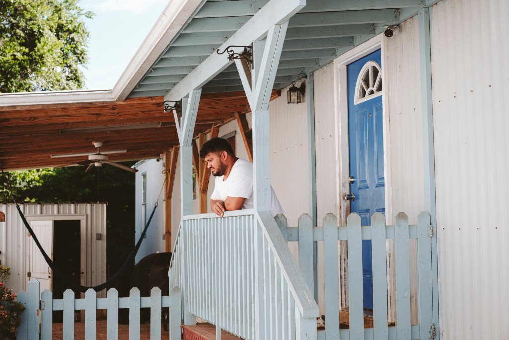 At the center of the image, a man stands leaning on a stairway railing, his gaze cast downward. Behind him and to the left is a covered patio with a hammock, a ceiling fan, and some chairs. To the right is a brightly-colored door to a trailer home.