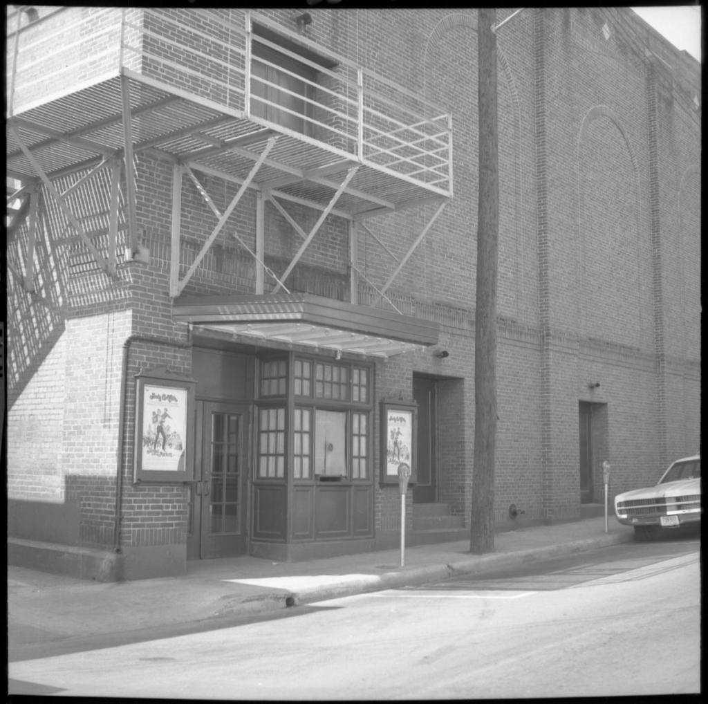 A black and white image of a brick building with back doors and theater posters, and a metal railing to a second story entrance.
