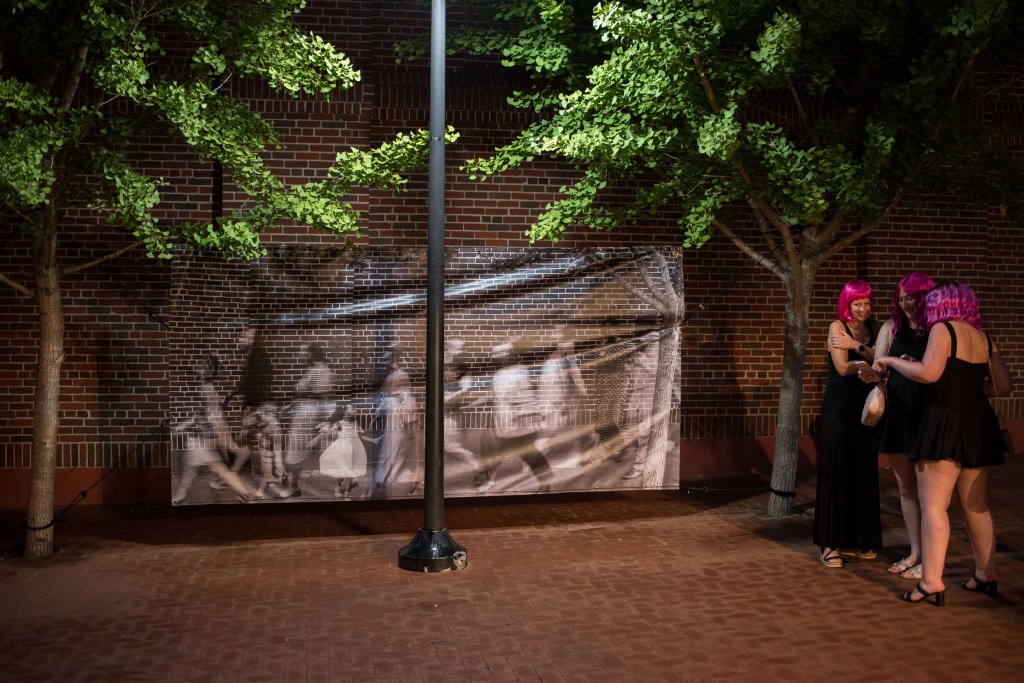 Three people in black dresses and pink wigs on a brick lined street, to the side of a large format black and white image, with trees to the side.