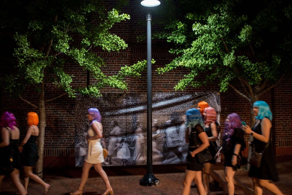 A group of people in colorful wigs, all in black dresses except one in white, walk past a large format, black and white print, with trees on the sides.