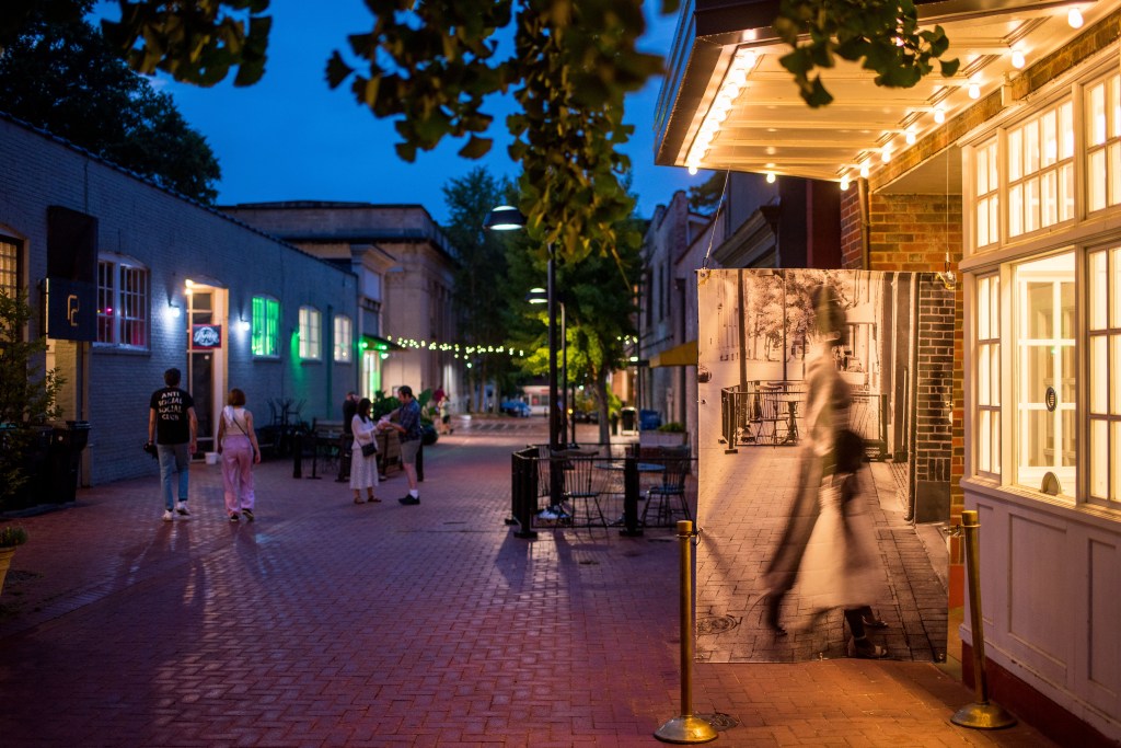 A brick lined road with buildings on either side and people walking through. On the side is a large format black and white image of a life-size person walking, in positing toward the actual box office.