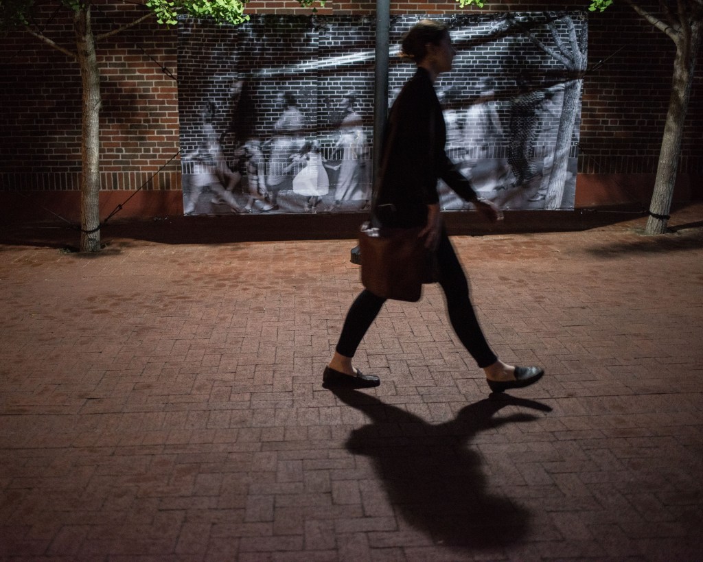 A person in black, with a shoulder bag, walking by a large format black and white image on a brick lined road.