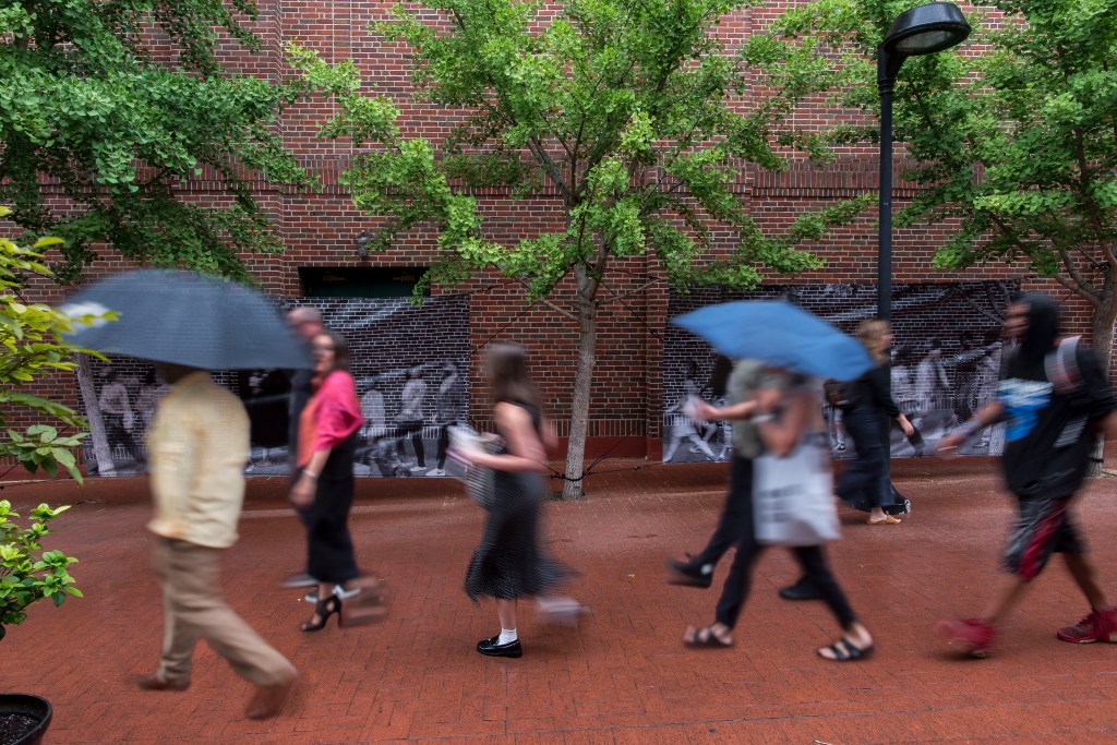 People walk on a brick lined road, a bit blurry, with two large black and white panels behind them and trees in between.