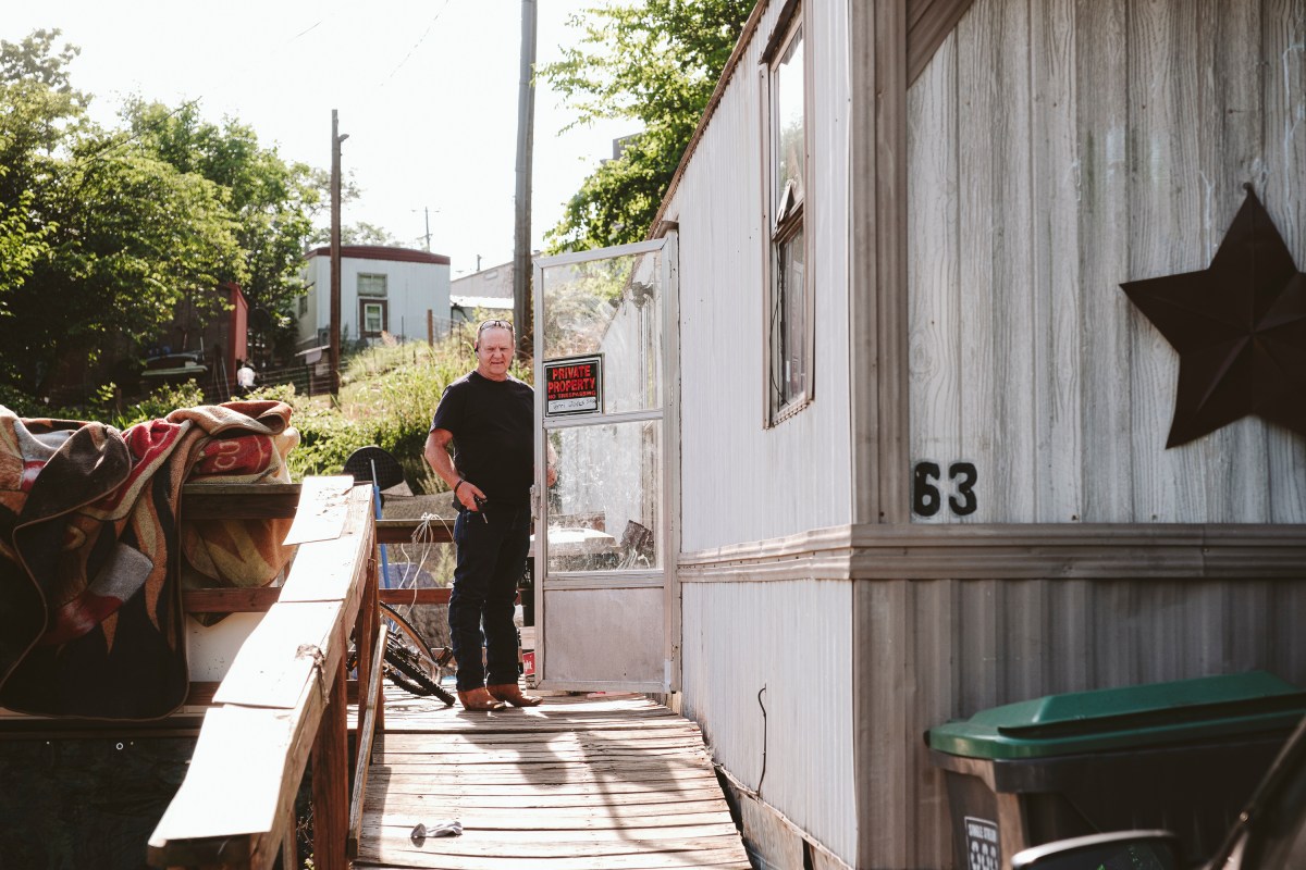 A man in cowboy boots, jeans, and a t-shirt looks at the camera as he opens a screen door. He is standing on a wooden deck built alongside a trailer home. On the door is a "private property" sign. On the side of the trailer, close to the camera, is a decorative metal star.