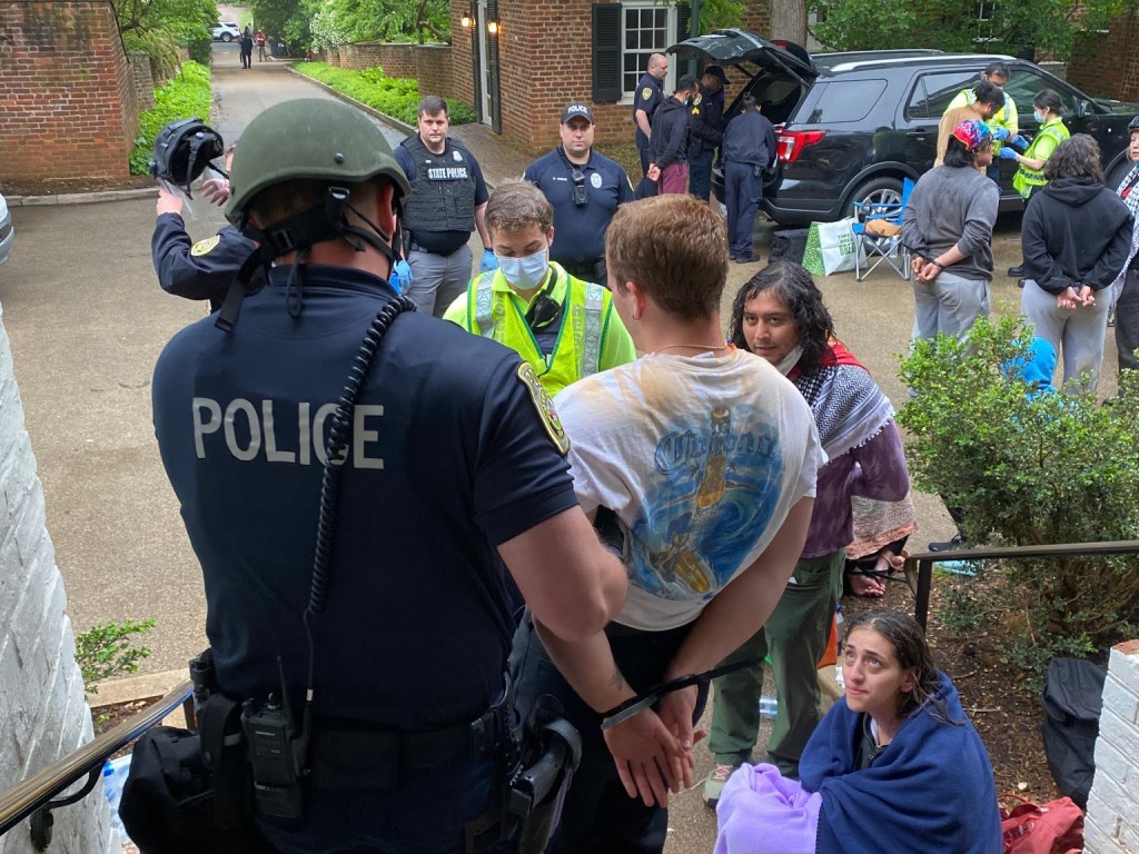 Police officer lead a person in a t-shirt, hands tied behind his back, dow a stairwell, while others look on.