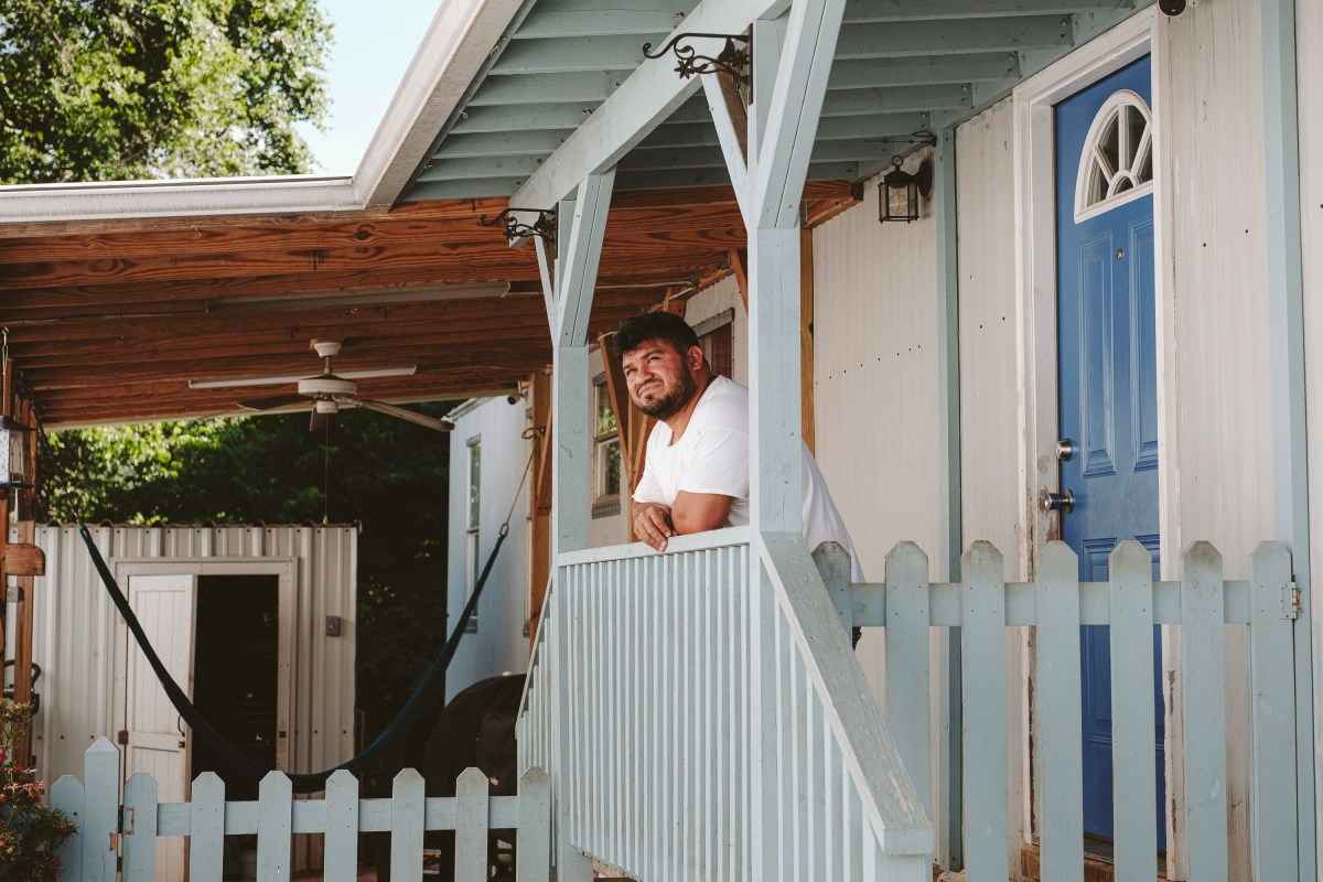 At the center of the image, a man stands leaning on a stairway railing, his gaze cast upward. Behind him and to the left is a covered patio with a hammock, a ceiling fan, and some chairs. To the right is a brightly-colored door to a trailer home.