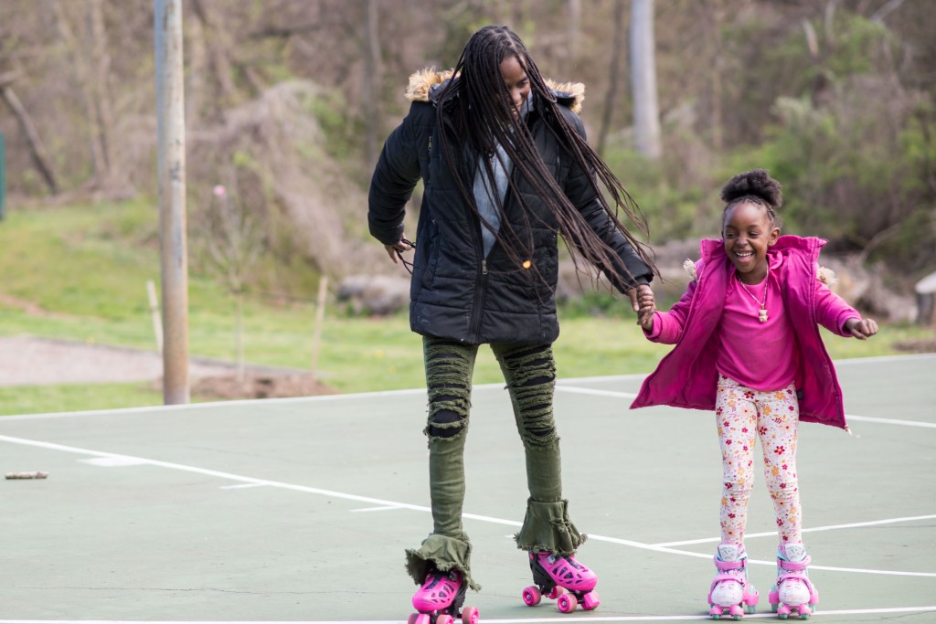 A woman and child hold hands while rollerstaking on an outdoor basketball court. Both are smiling and laughing.