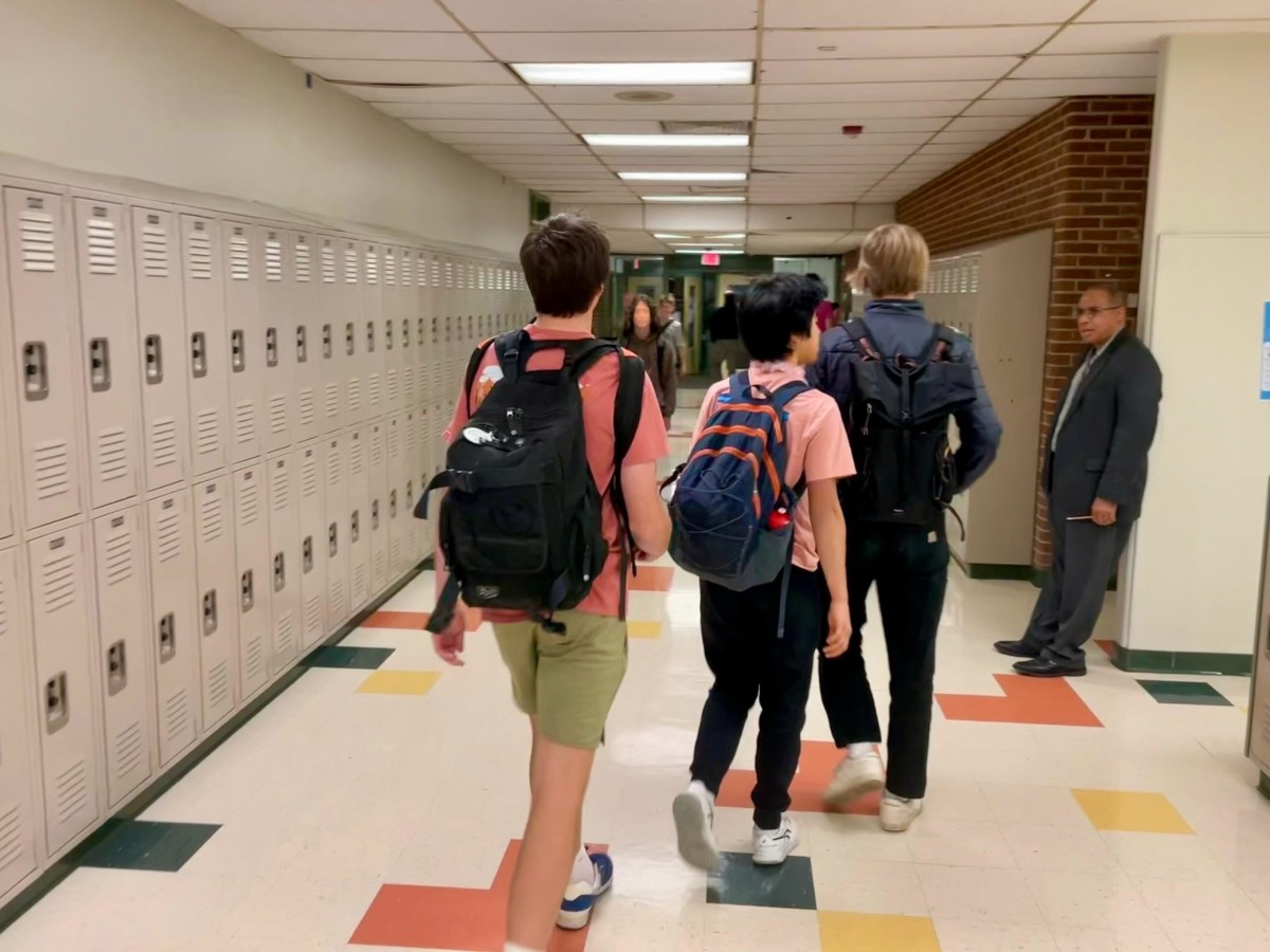 Teens wearing backpacks walk down a school hallway beside a row of lockers.
