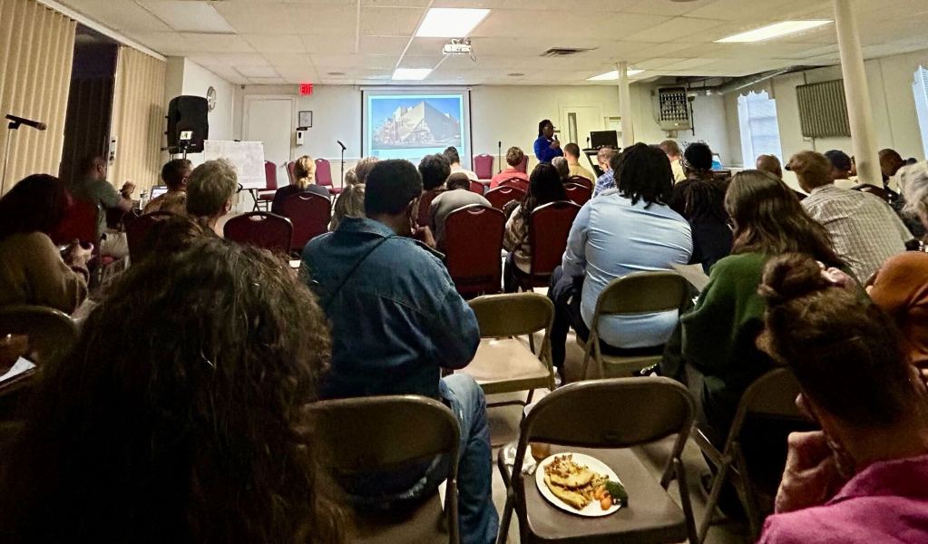 People sit in rows of chairs in a narrow room with low ceilings. They face a projection screen that shows an architect's rendering of a building. People are taking notes and eating pizza.