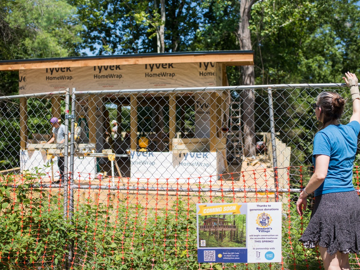 A woman stands at a chainlink fence, looking at a mostly completed structure surrounded by trees.