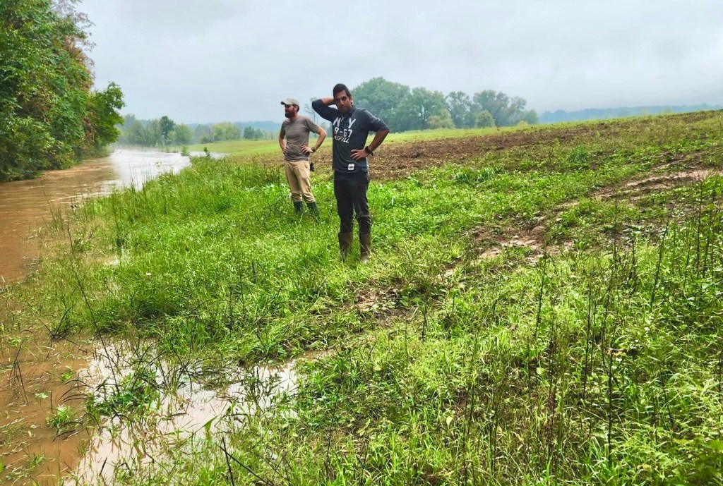 Two men stand in next to a submerged agricultural field looking helplessly on.