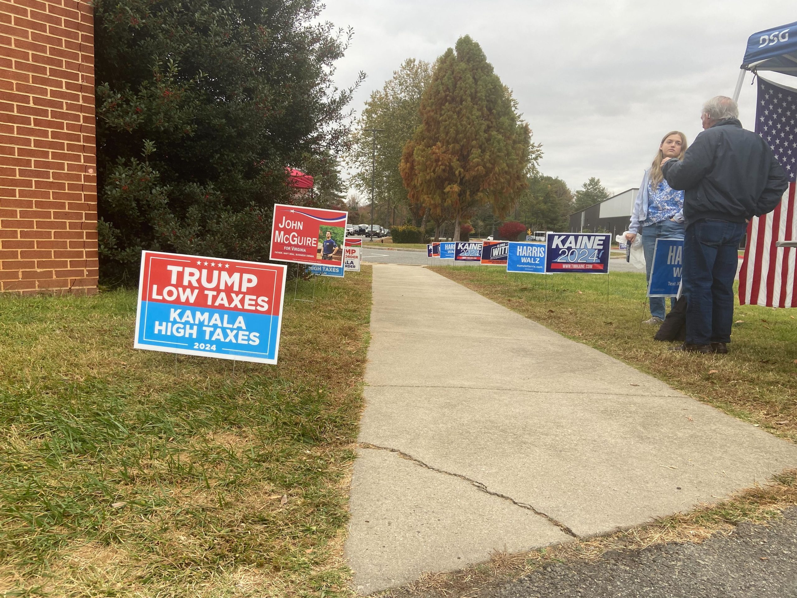 A low view of signs on either side of a walkway next to a brick building. On one side are mostly red signs for Trump and McGuire; on the other, mostly blue for Harris and Kaine.
