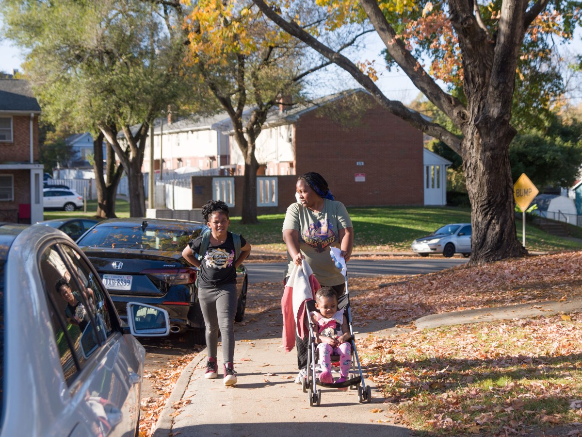 A woman pushes a stroller down a leafy sidewalk, with a young child seated another child wearing a backpack walking beside them. There are two storey brick buildings behind them.