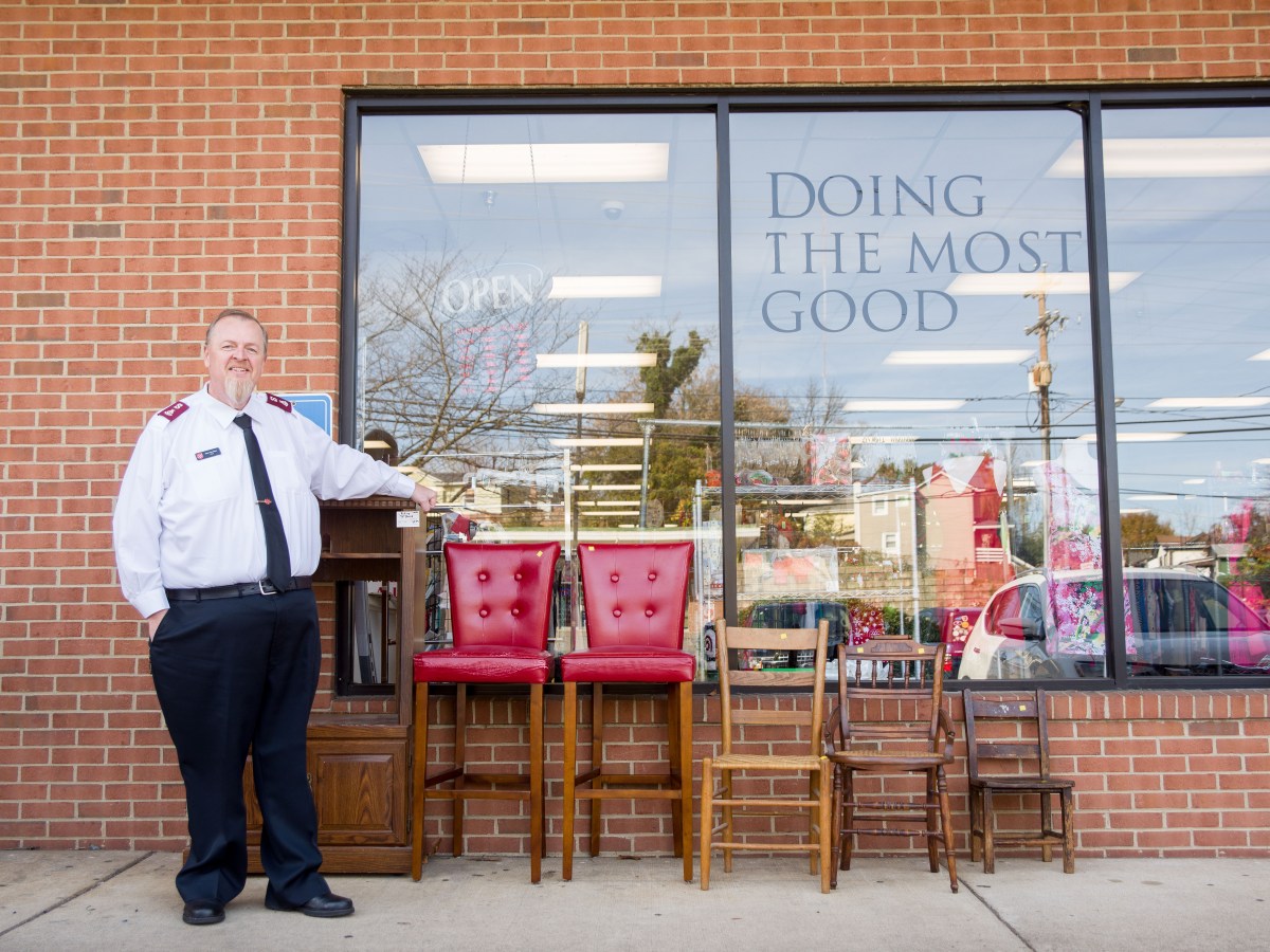 A man stands in front of a brick building with three plate glass windows. He leans on a cabinet, and next to him is a line of five chairs of varying sizes. One of the windows has a decal that says, "Doing the most good."